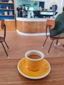 Yellow cup of espresso on a wooden table with the coffee bar behind at Sukhum Caffe Sanpatong