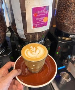 Flat white with latte art held in front of the bean hoppers at The Larder Cafe & Bar