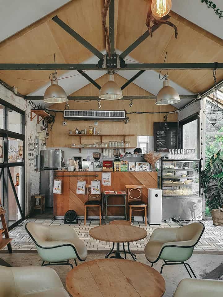 The Barn's coffee bar under a pitched plywood ceiling, with a wooden counter, pastry cabinet and pale-green armchairs on round wooden tables