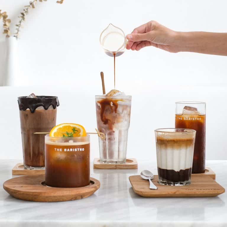 A hand pouring syrup over an array of The Baristro specialty iced coffee drinks garnished with orange